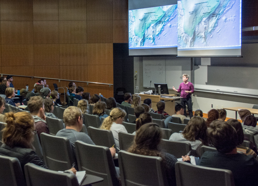 A lecture hall full of people with a presentation featuring topographical maps projected behind the speaker