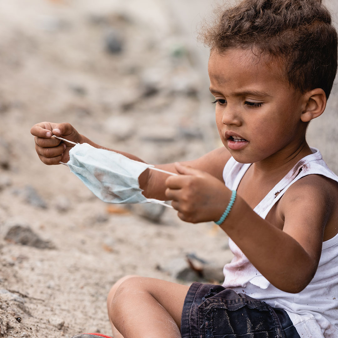 A young child sitting on a beach holding a disposable face mask surrounded by pollution